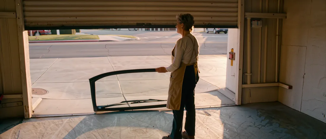 A Raquels W House technician in a work apron carrying a replacement windshield in through the open roll-up bay door